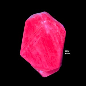 Könnte beinhalten: Ein leuchtend rosa Kristall mit strukturierter Oberfläche, vor schwarzem Hintergrund. Der Kristall hat eine einzigartige, unregelmäßige Form und die Worte "Long wave" sind sichtbar.