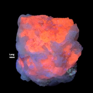 May include: A close-up of a mineral specimen with a vibrant orange glow emanating from within. The outer layer has a light blue hue with hints of purple and blue specks. The words "Long wave" are visible on the left side.