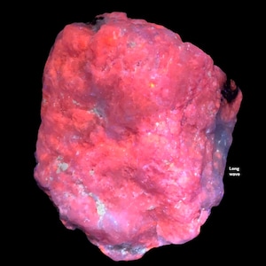 May include: A close-up of a glowing, rough-textured rock with a vibrant pink hue. The rock has a complex, uneven surface with hints of blue and white. The words "Long wave" are visible on the right side of the image against a black background.