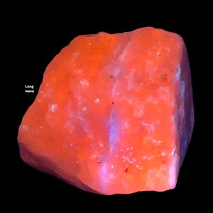 May include: A vibrant orange mineral specimen with a rough, textured surface. The rock has a glowing appearance, with hints of blue and purple. The words "Long wave" are visible in the image. The background is black.