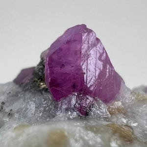 Könnte beinhalten: Nahaufnahme einer lebendigen, facettierten, rosa Kristallformation. Der Kristall ist der Mittelpunkt, vor dem Hintergrund einer helleren, strukturierten mineralischen Basis. Die Oberfläche des Kristalls reflektiert das Licht und zeigt seine natürliche Struktur.