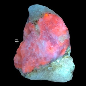 May include: A glowing, irregular-shaped mineral specimen with a vibrant red and pink core, surrounded by a pale blue-green exterior. The text "Long wave" is visible on the left side. The background is black.