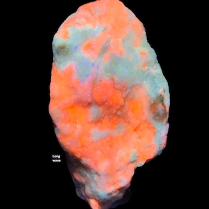 May include: A glowing, irregular-shaped mineral specimen. The rock has a vibrant orange hue with patches of pale green and hints of blue. The words "Long wave" are visible in the lower left corner against a black background.