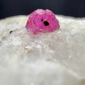 Könnte beinhalten: Nahaufnahme eines leuchtend rosa Rubinkristalls mit dunklem Einschluss, vor dem Hintergrund von weißem, kristallinem Gestein. Die Oberfläche des Rubins ist strukturiert und reflektiert das Licht, wodurch seine natürliche Schönheit zur Geltung kommt.
