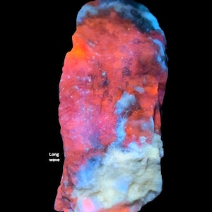 May include: A glowing rock specimen with a black background. The rock displays vibrant colors, including red, orange, blue, and white, with the text "Long wave" visible. The colors suggest fluorescence under long-wave UV light.