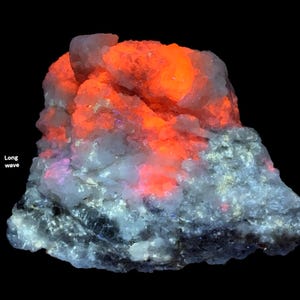 May include: A close-up of a mineral specimen with a vibrant orange glow emanating from the top, contrasted by a gray and white base. The text "Long wave" is visible on the left side. The mineral appears to be illuminated by a specific light source.