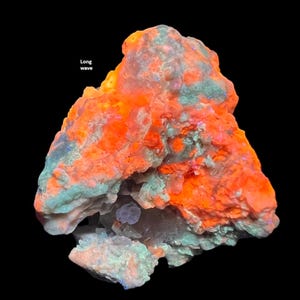 May include: A fluorescent mineral specimen with vibrant orange and green hues against a black background. The text "Long wave" is visible in the upper left corner. The mineral's surface has a rough, textured appearance.