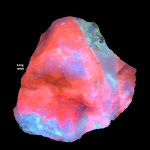 May include: A glowing, rough-hewn mineral specimen with a vibrant color palette. The rock displays a striking combination of red, pink, and blue hues, with the text "Long wave" visible. The colors are most intense on the surface of the rock.