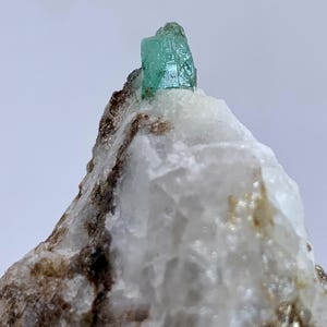 Puede incluir: Primer plano de un espécimen mineral con una vibrante formación de cristal verde claro sobre una matriz de roca blanca y marrón. El cristal tiene una superficie texturizada y la roca tiene un aspecto natural rugoso. El fondo es de color neutro.