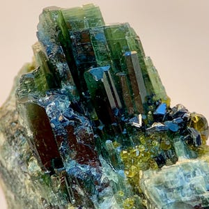 Op de afbeelding: Een close-up van een mineraalmonster met een cluster van groene, blauwe en bruine kristallen. De kristallen hebben een prismatische structuur en zijn in een gelaagde formatie gerangschikt. Kleine, geelgroene kristallen zijn ook zichtbaar.