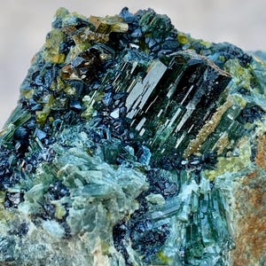 Könnte beinhalten: Nahaufnahme einer Mineralprobe mit einem Cluster aus länglichen, durchscheinenden grünen Kristallen. Die Kristalle sind mit dunkelblauen und goldbraunen Formationen durchsetzt, wodurch ein strukturiertes, natürliches Aussehen entsteht. Die Farbpalette ist eine Mischung aus Grün-, Blau- und Brauntönen.