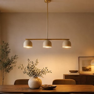 May include: A linear pendant light fitting featuring a slim brass-toned rod suspended from the ceiling, supporting three downward-facing dome-shaped shades in a matte cream finish, each emitting a warm glow from the integrated light source.