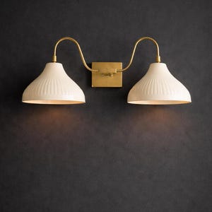 May include: A double wall light with a brass finish and cream, ribbed shades. The fixture has a rectangular base and curved arms. The sconce is mounted on a dark grey wall, providing a contrast to the warm tones of the light.