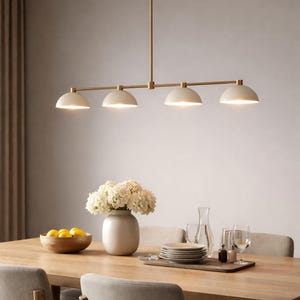 May include: A modern dining room setting featuring a wooden table, chairs, and a four-light pendant lamp. The lamp has a brass finish and cream-colored dome shades. A vase of white flowers and a bowl of lemons are on the table.