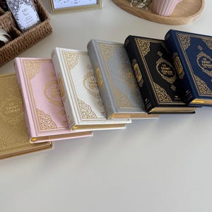 Puede incluir: Una colección de libros de tapa dura, cada uno con letras doradas y detalles dorados decorativos. Los libros son de varios colores, incluyendo dorado, rosa, blanco, gris, azul marino y azul. El texto "LE SAINT CORAN" es visible.