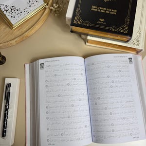 Peut inclure: Un livre ouvert avec du texte arabe, un stylo noir et argenté dans une boîte blanche et une pile de livres avec des détails dorés. Le livre a des bords roses et est ouvert sur une page avec une écriture arabe. Une image encadrée et des fleurs séchées sont également visibles.