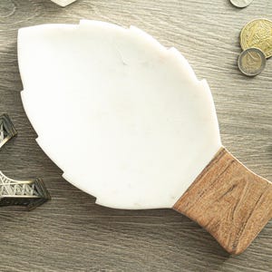 May include: A white marble leaf-shaped tray with a wooden handle. The tray is on a wooden surface, with a miniature Eiffel Tower and coins nearby. The tray's design is reminiscent of nature.