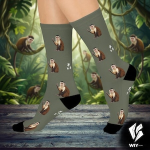 May include: Olive green socks featuring a repeating pattern of brown and white capuchin monkeys. The socks have black heels and toes, with white star accents. The background shows a jungle scene with more monkeys.