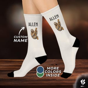 Könnte beinhalten: Ein Paar weiße Socken mit einer Illustration eines braunen Faultiers, das an einem Ast hängt, und dem in schwarzer Schrift gedruckten Namen ALLEN. Die Socken haben schwarze Fersen und Zehenbereiche. Das Bild enthält den Text CUSTOM NAME und MORE COLORS INSIDE.