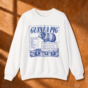 Puede incluir: Una sudadera blanca de manga larga con un estampado gráfico azul de una cobaya. El diseño incluye el texto GUINEA PIG, detalles científicos, puntos sobre el animal y una ilustración de tres cobayas en la parte inferior.