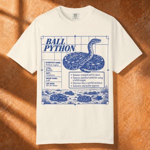 May include: An off-white short-sleeve t-shirt featuring a blue graphic print of a ball python. The design includes a grid background, the text BALL PYTHON, scientific details, and bullet points describing the snake's traits and habitat.