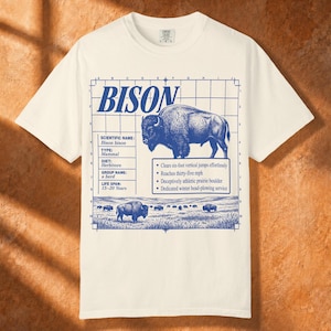 May include: An off-white t-shirt featuring a blue graphic print of a bison. The design includes a grid, the word BISON, scientific facts, and a landscape illustration. Text includes Scientific Name: Bison bison, Type: Mammal, Diet: Herbivore, Group Name: a herd, Life Span: 15-20 Years, and bullet points about bison traits.
