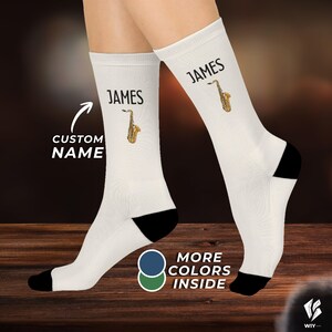 May include: A pair of white crew socks featuring a black heel and toe, with the name JAMES printed in black above a small graphic of a gold saxophone. The text CUSTOM NAME with an arrow and MORE COLORS INSIDE are also visible on the image.