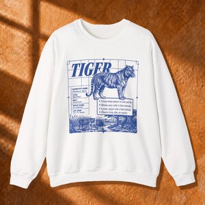 Puede incluir: Una sudadera blanca de manga larga con un estampado gráfico azul de un tigre. El diseño incluye la palabra TIGER, detalles científicos y datos sobre el animal, sobre un fondo de cuadrícula con una escena de bosque en la parte inferior.