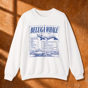 Pode incluir: Uma camisola branca de manga comprida com uma impressão gráfica azul de uma beluga. O design inclui o texto BELUGA WHALE, detalhes científicos, uma lista de factos sobre o mamífero e uma ilustração da baleia na água.