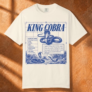 Puede incluir: Una camiseta de manga corta color hueso con un estampado gráfico azul de una cobra real. El diseño incluye el texto KING COBRA, detalles científicos y puntos sobre el comportamiento de la serpiente dentro de un marco con forma de cuadrícula.