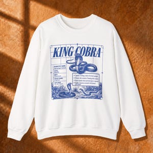 Puede incluir: Una sudadera blanca de manga larga con un estampado gráfico azul de una cobra real. El diseño incluye una cuadrícula, datos científicos e ilustraciones de serpientes en su hábitat natural. El texto dice KING COBRA y otros detalles.