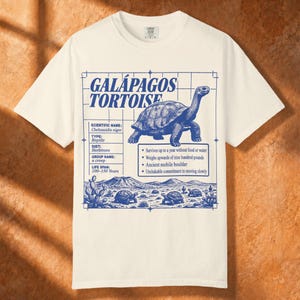 Peut inclure: Un t-shirt couleur crème avec une impression graphique bleue représentant une tortue des Galápagos. Le motif inclut des faits scientifiques, un arrière-plan quadrillé et un paysage désertique avec plusieurs tortues et cactus en style trait.