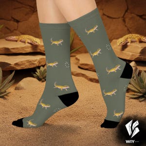 May include: Olive green socks featuring a repeating pattern of yellow and black geckos and white starbursts. The socks have black heels and toes. The socks are shown on a person's legs.