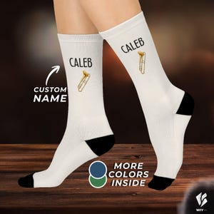 Op de afbeelding: Een paar witte sokken met een zwarte hiel en teen. Elke sok toont de naam CALEB in zwarte tekst boven een kleine afbeelding van een gouden trombone. De afbeelding bevat de tekst CUSTOM NAME en MORE COLORS INSIDE.