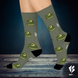 Puede incluir: Calcetines verde oliva con un patrón repetido de diseños en espiral verdes, amarillos y blancos. Los calcetines tienen talones y dedos negros, y líneas onduladas blancas. Los calcetines se muestran en las piernas de una persona.
