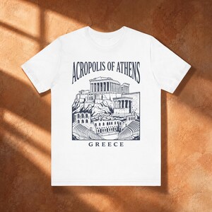 May include: A white short-sleeved t-shirt featuring a dark blue graphic print of the Acropolis of Athens. The design includes detailed illustrations of ancient Greek ruins and the text Acropolis of Athens Greece centered on the chest area.