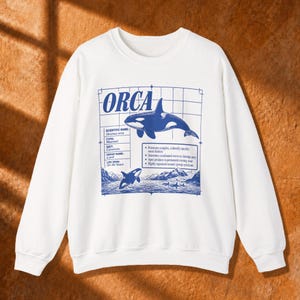 Può includere: Una felpa bianca a maniche lunghe con una stampa grafica blu di un'orca. Il design include il testo ORCA, dettagli scientifici e uno sfondo a griglia. L'illustrazione raffigura orche nell'oceano con montagne in lontananza.