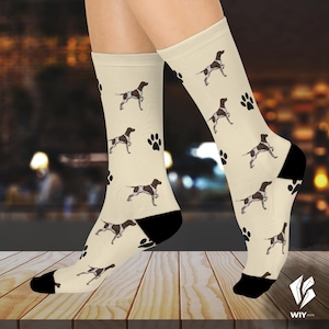 Może przedstawiać: Kremowe skarpetki z powtarzającym się wzorem brązowo-białych psów i czarnych odcisków łap. Skarpetki mają czarne pięty i palce. Skarpetki znajdują się na drewnianej powierzchni.