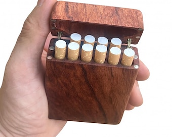 Puede incluir: Un estuche de cigarrillos de madera, abierto para revelar varios cigarrillos. El estuche es de madera marrón oscuro con una tapa con bisagras. Los cigarrillos tienen filtros blancos y papel marrón claro.