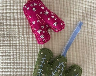 May include: Two felt Christmas ornaments. One is a magenta letter "A" with white snowflake embroidery and clear gem accents, and a pink ribbon. The other is a green letter "M" with white embroidered branches and clear gem accents, and a blue ribbon.