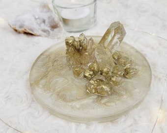 May include: A clear glass dish with a gold glitter finish and a cluster of clear quartz crystals embedded in the center. The dish is sitting on a white fluffy surface.