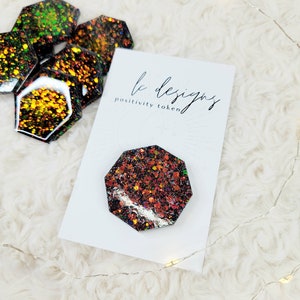 May include: Glittery, multi-colored positivity tokens with a black base and a white card that reads "le designs positivity token". The tokens are octagonal and have a shiny, textured appearance. The background is a soft, white, fluffy surface.