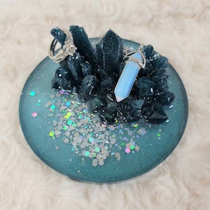May include: A teal blue ring dish with iridescent glitter and a black crystal cluster. A silver ring and a silver pendant with a light blue crystal are displayed on the dish.