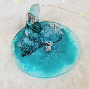 May include: A turquoise blue resin ring dish with a crystal-like design. The dish features two silver rings and a silver ring with a monogram.