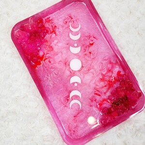 May include: Pink resin tray with a white moon phase design. The tray has a swirled, marbled effect and pink glitter embedded in the resin.