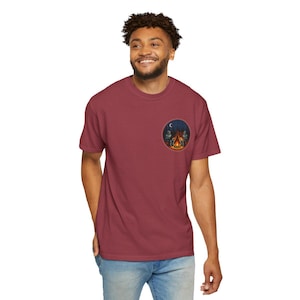May include: Burgundy t-shirt with a circular graphic depicting a campfire scene under a crescent moon. The graphic includes trees and figures around the fire. The t-shirt is made of a soft material, suitable for casual wear.