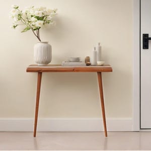 May include: A mid-century modern style wooden console table with a light brown finish and tapered legs. The table is adorned with a white vase holding white flowers, along with decorative bottles, small bowls, and stacked plates. The table is 30 inches long.