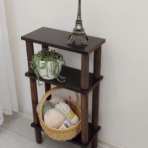 Puede incluir: Estantería de madera marrón oscuro de tres niveles. La balda superior contiene una pequeña figura de la Torre Eiffel y una maceta blanca con una planta verde. La balda intermedia tiene una cesta tejida llena de hilo. La unidad está sobre un suelo claro.