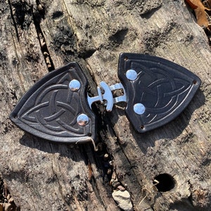 May include: A black leather belt buckle with a Celtic knot design and silver accents. The buckle is made of two pieces that snap together.