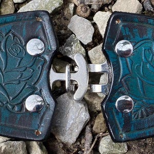 Pu&ograve; includere: Un paio di fibbie per cintura in pelle blu turchese con un design a rosa e chiusure in metallo argentato.